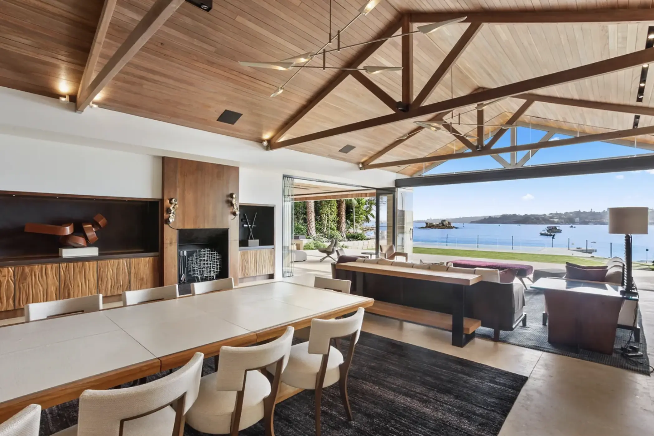 Luxurious open-plan dining and living area with vaulted wooden ceilings and exposed beams, seamlessly extending to an outdoor patio with stunning waterfront views. A large dining table with upholstered chairs sits in the foreground, while modern sofas and lounge furniture face the expansive glass doors that open to the ocean. Natural light floods the space, highlighting the contemporary décor, wooden accents, and a stylish fireplace.