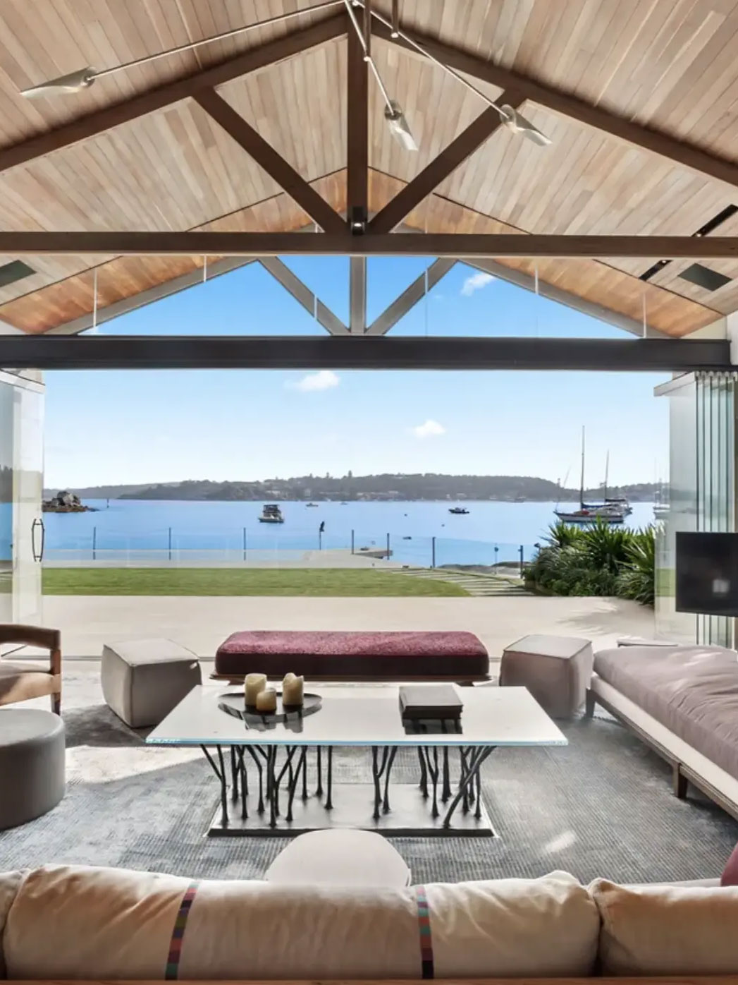 vaucluse_house_living_ Luxury living room at Vaucluse Estate, designed by Nina Maya Interiors. The open-plan space features a vaulted timber ceiling with exposed beams, plush modern furnishings, and a statement glass coffee table. Floor-to-ceiling glass doors offer a seamless indoor-outdoor flow, showcasing breathtaking waterfront views with yachts and lush greenery in the background.